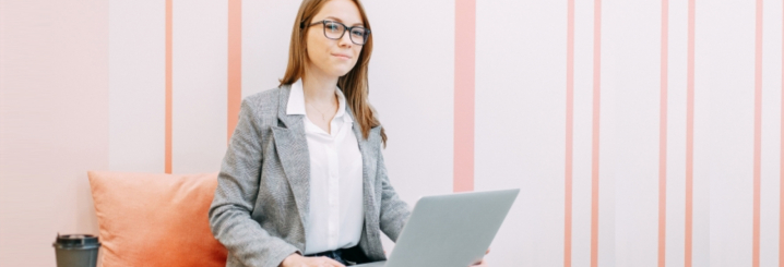 Junge Frau mit Brille und Laptop sitzt auf einem Sofa mit Kissen, neben ihr steht ein Coffee-to-go- - TOP-Kurs